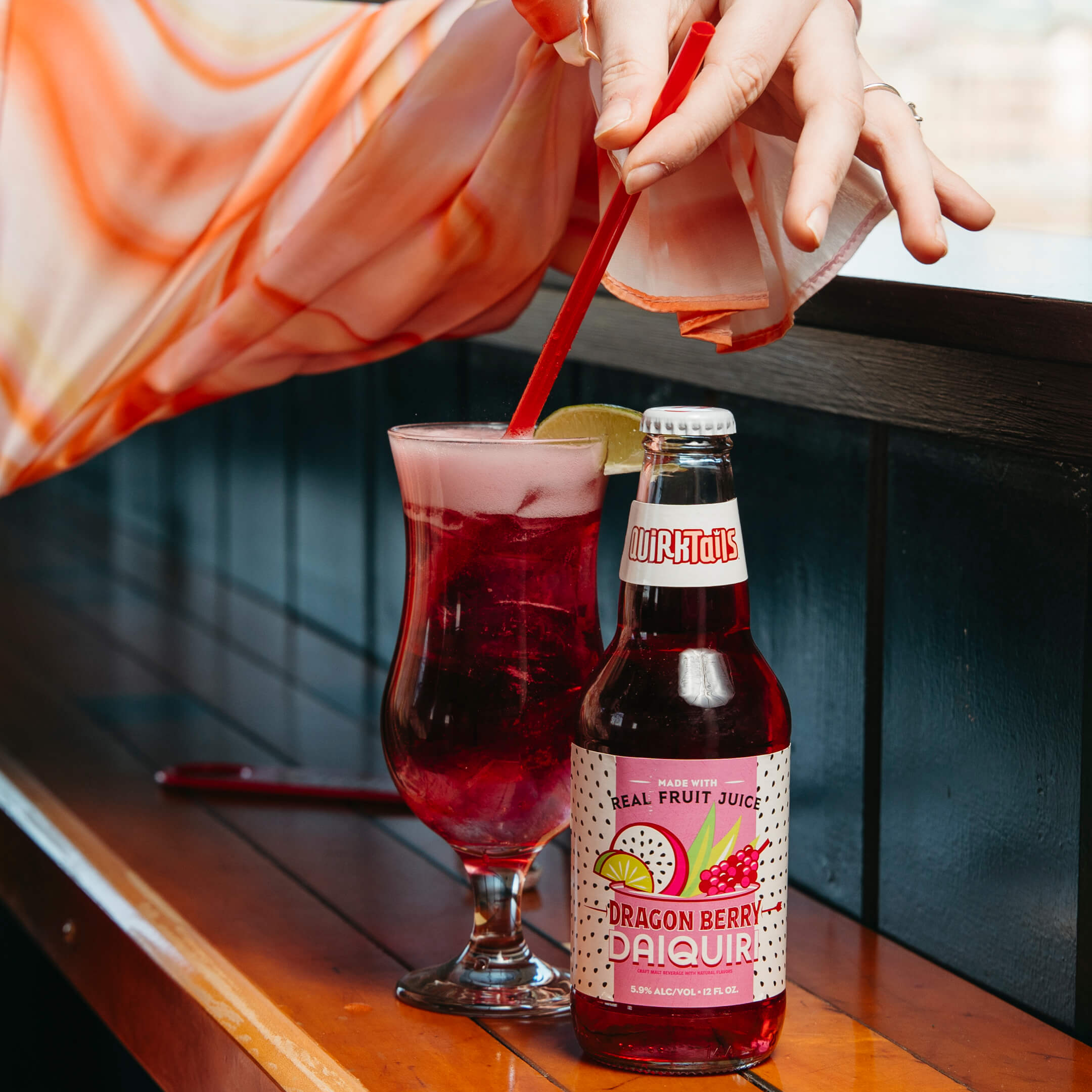 A hand holds a straw in a glass filled with a pink drink topped with foam and a lime slice. Beside it is a bottle labeled "Dragon Berry Daiquiri." Both are placed on a wooden surface with a blurred background.