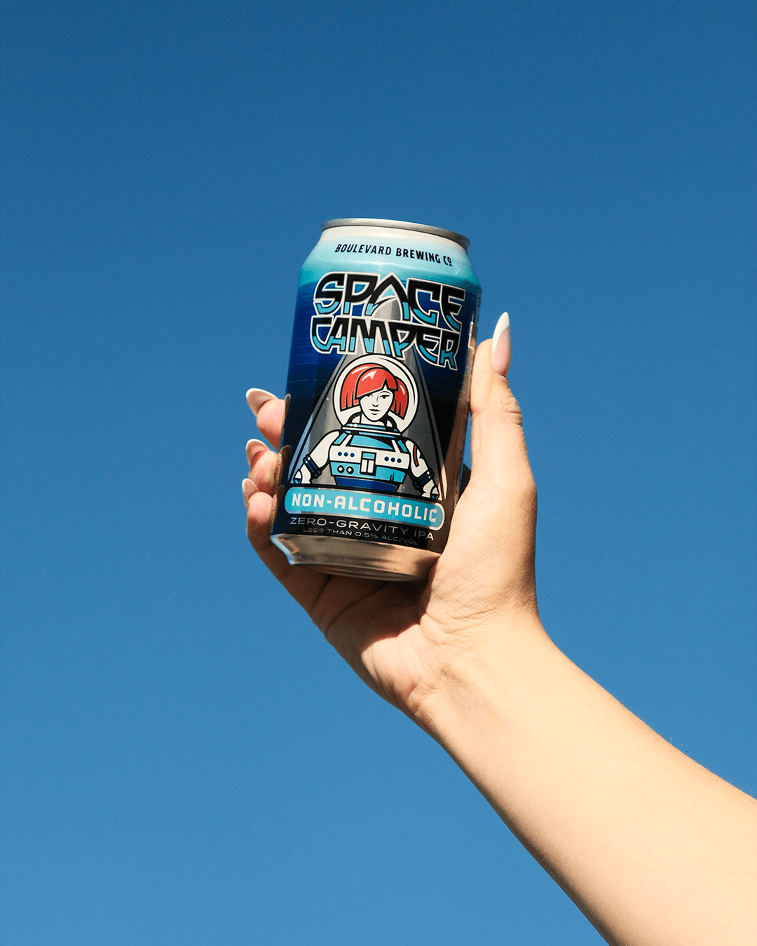 A hand with manicured nails holds up a can of non-alcoholic "Space Camper" beverage against a clear blue sky. The can features a cartoon astronaut in blue and white. The text reads "Boulevard Brewing Co." and "Zero-Gravity.