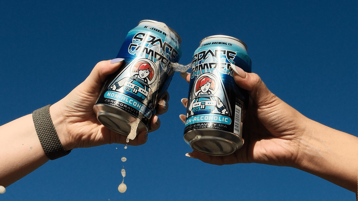 Two hands clink together cans of "Space Camper" non-alcoholic beer against a clear blue sky. Foam spills from the cans, each featuring a cartoon space-themed design on the label.