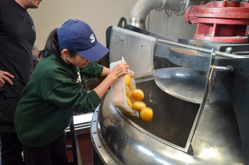 A person wearing a blue cap and green sweater is adding oranges into a large industrial machine, observed by another person. The scene appears to be in a factory setting with stainless steel equipment.