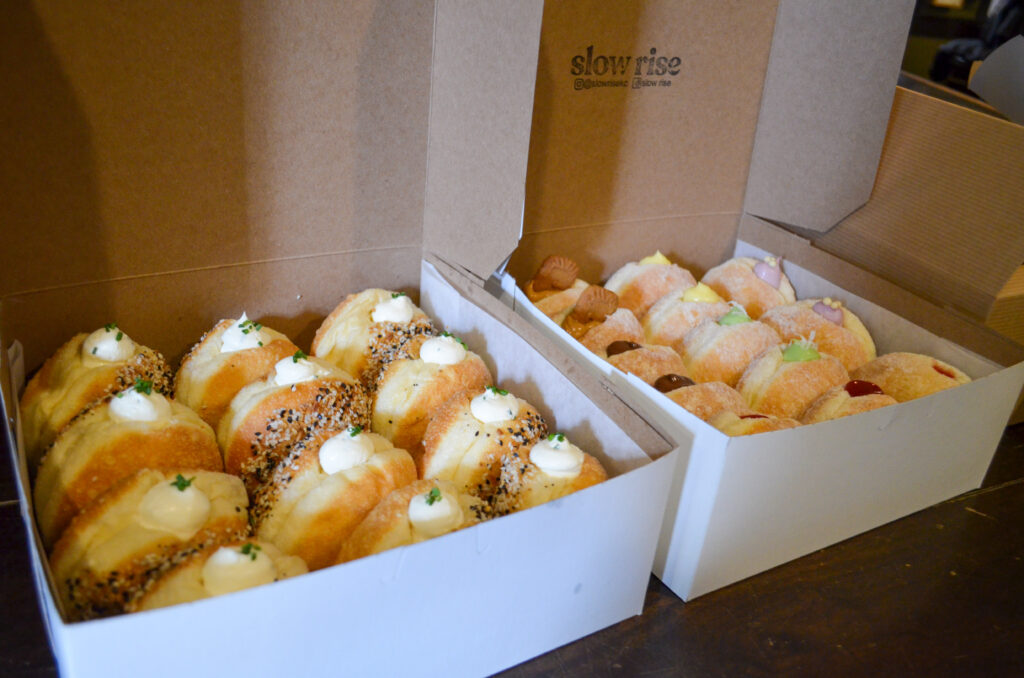 Two open boxes filled with assorted pastries on a wooden table. The left box has pastries topped with cream and herbs, sprinkled with seeds. The right box contains smaller pastries, some topped with powdered sugar or jam.