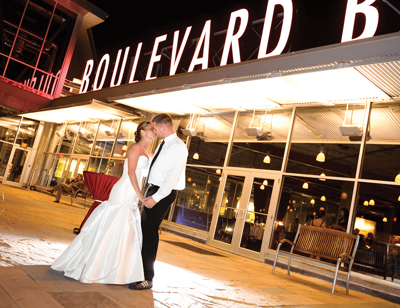 A couple dressed in wedding attire dances beneath a large "Boulevard" sign outside a modern, well-lit building. The romantic scene, set against reflections on the glass windows behind them, captures the enchanting allure of Boulevard Rental Spaces.