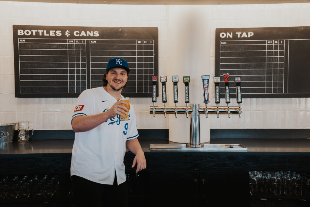 Vinnie Pasquantino in a baseball jersey and cap holds a beer glass, smiling in front of a beer tap setup and menu board.